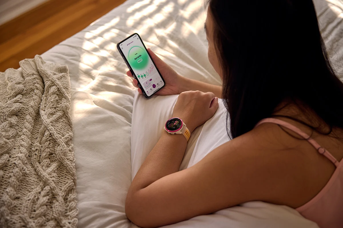 Kvinde, der ligger i sengen og bruger Natural Cycles° appen på mobilen med et Garmin smartwatch på håndleddet.